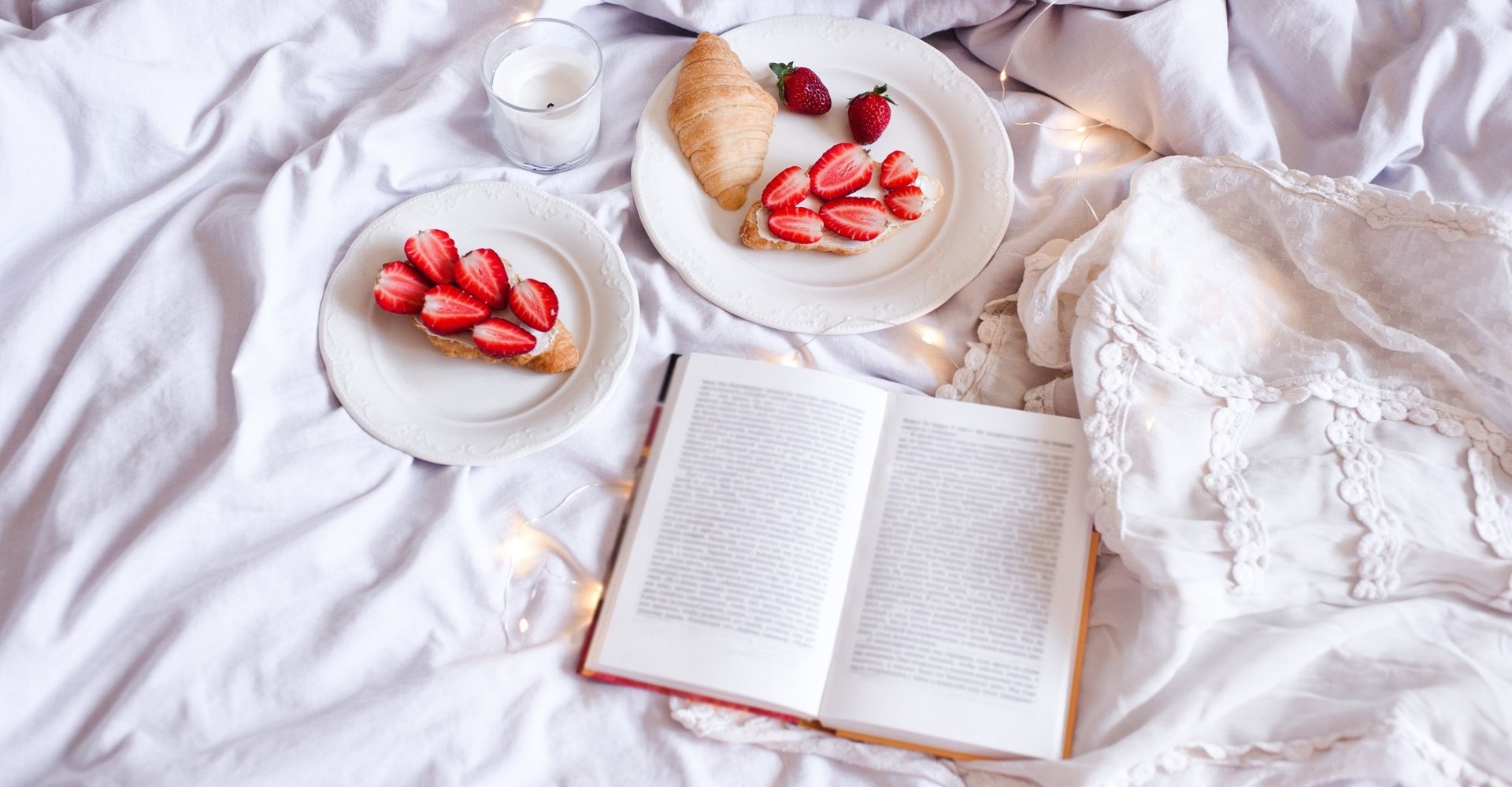 Un libro aperto sul lenzuolo bianco di un letto disfatto, con due piatti da colazione contenenti delle fragole