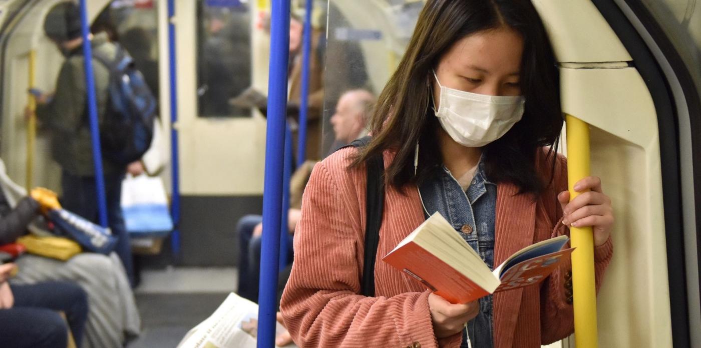 Una donna dai tratti orientali con addosso una mascherina legge in piedi su un mezzo pubblico un libro cartaceo