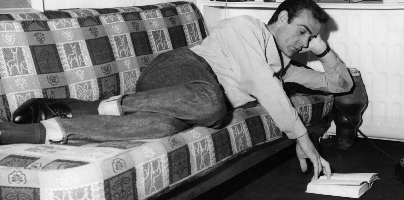 Sean Connery sdraiato su un divano legge un libro cartaceo appoggiato sul pavimento