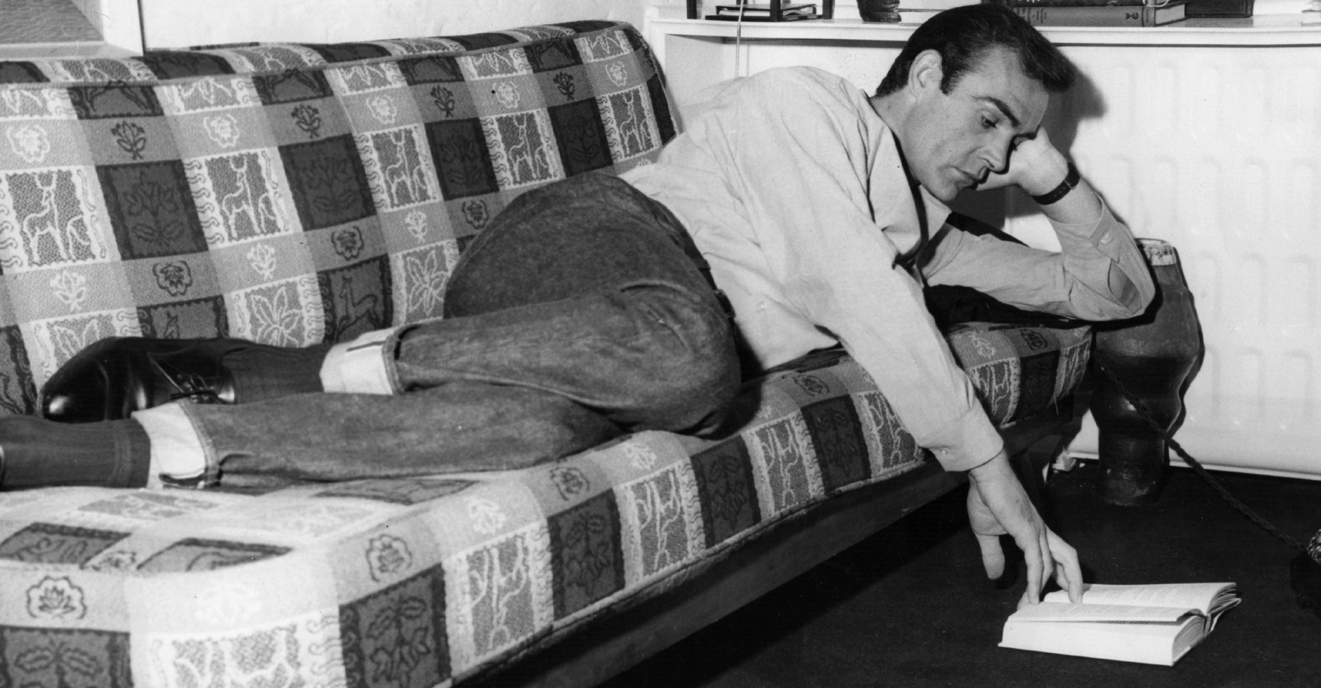Sean Connery sdraiato su un divano legge un libro cartaceo appoggiato sul pavimento