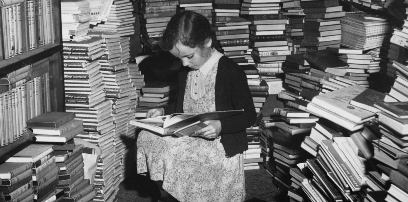 Foto in bianco e nero di una bambina che legge un libro seduta in una stanza piena di pile di altri libri,