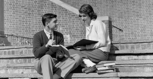 Due ragazzi seduti sui gradini di una scuola parlano fra di loro mentre leggono