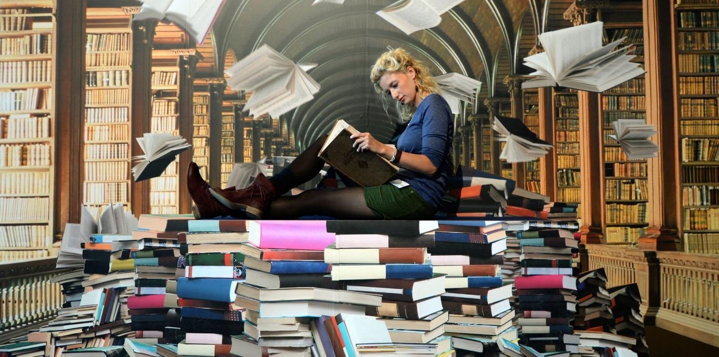 Una ragazza legge un libro con dietro lo sfondo di una biblioteca e sotto di sé l'installazione di diverse pile di libri