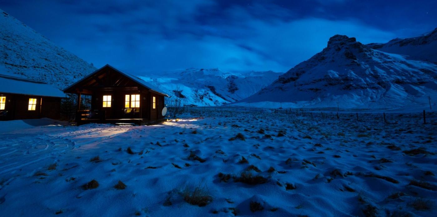 Immagine invernale di una casa di legno al chiaro di luna, con luce dalla finestra e montagne sullo sfondo, in Islanda meridionale
