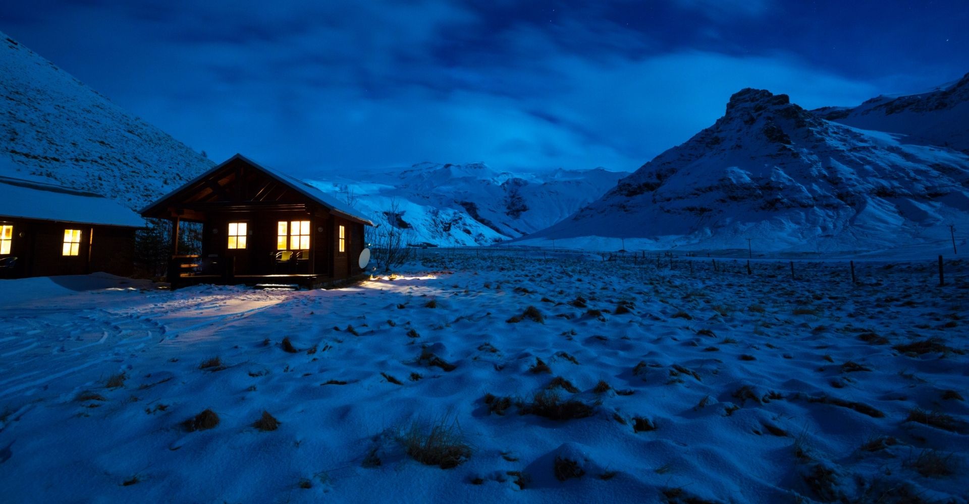 Immagine invernale di una casa di legno al chiaro di luna, con luce dalla finestra e montagne sullo sfondo, in Islanda meridionale