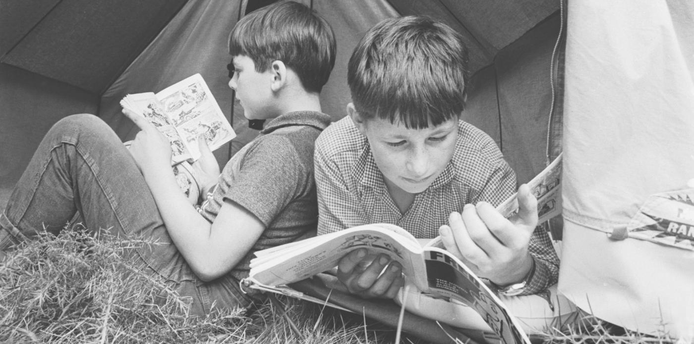 Foto vintage di due bambini che leggono dei fumetti sdraiati su un prato davanti a una tenda da campeggio