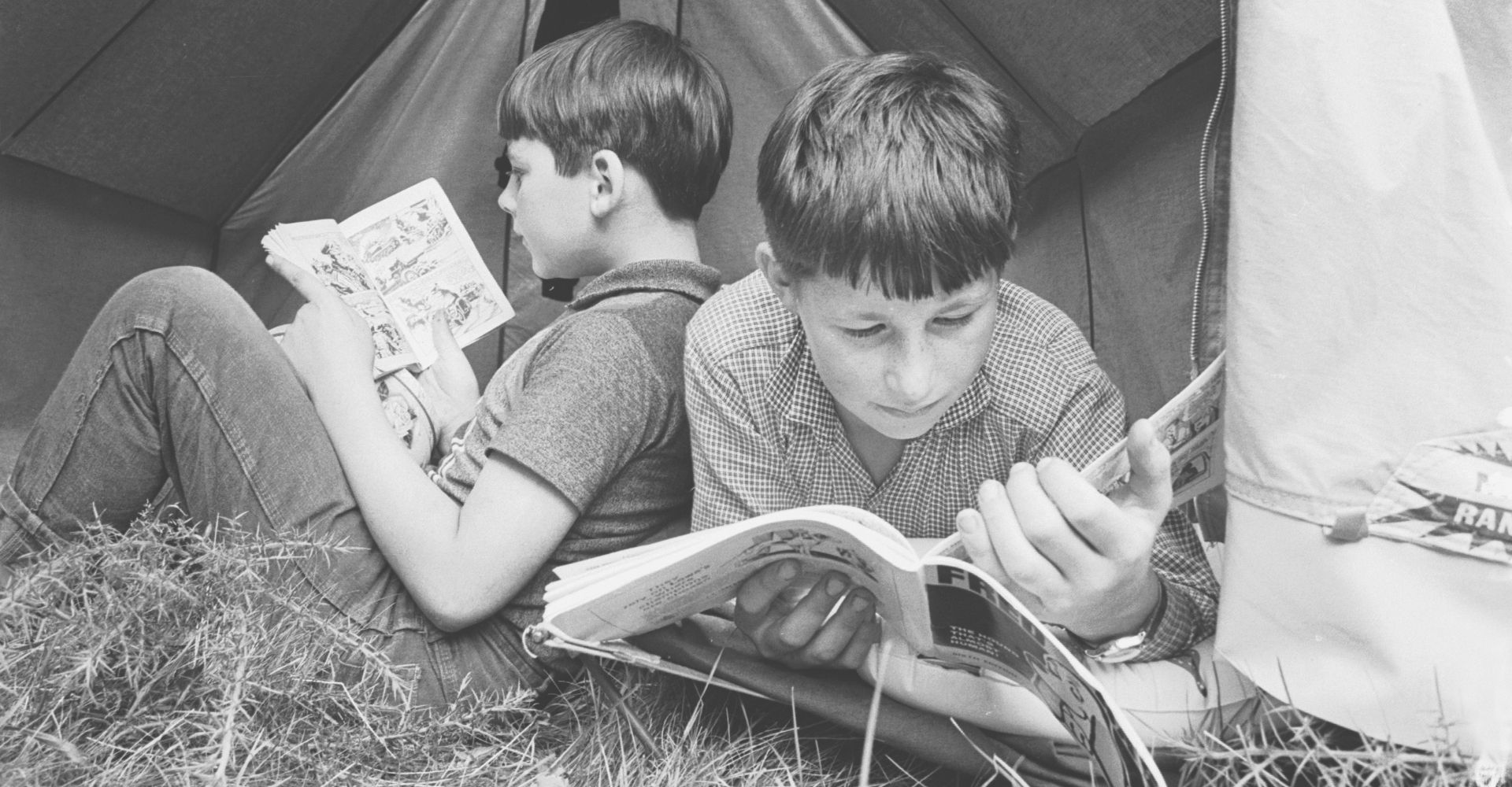 Foto vintage di due bambini che leggono dei fumetti sdraiati su un prato davanti a una tenda da campeggio