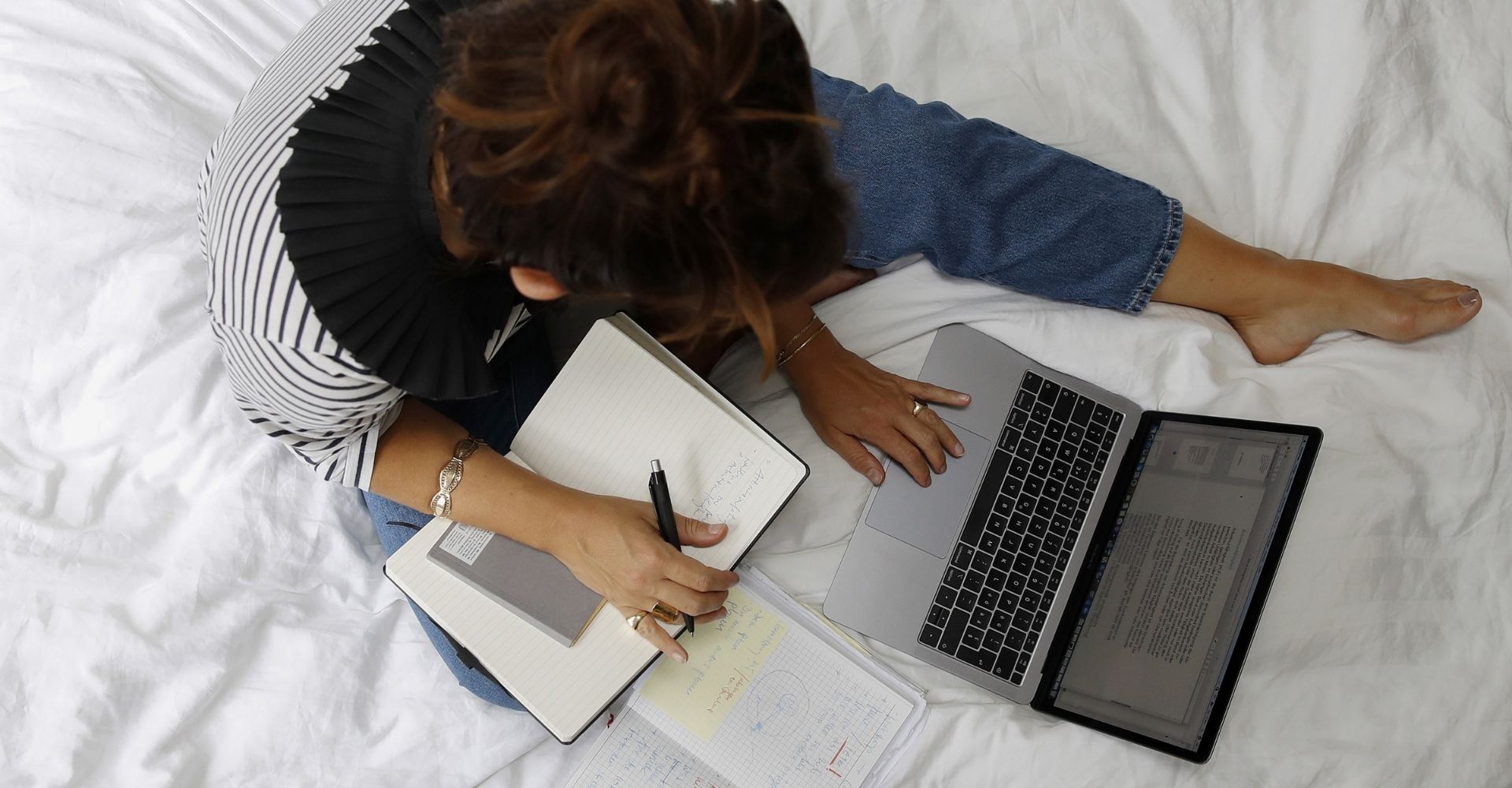 Vista dall'alto di una ragazza seduta su un letto, con un taccuino e dei fogli da una parte e un notebook dall'altra parte