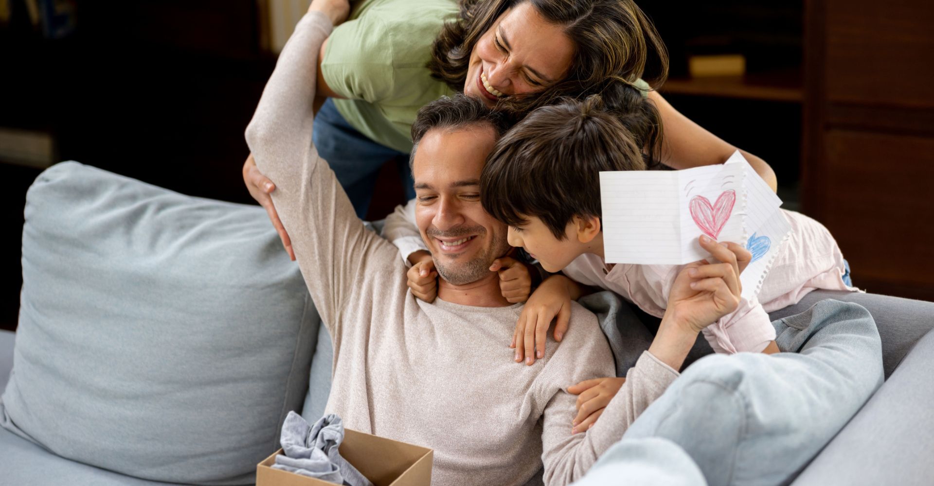 Un papà apre un regalo e legge un biglietto ricevuti per la Festa del papà mentre viene abbracciato da sua moglie e da suo figlio