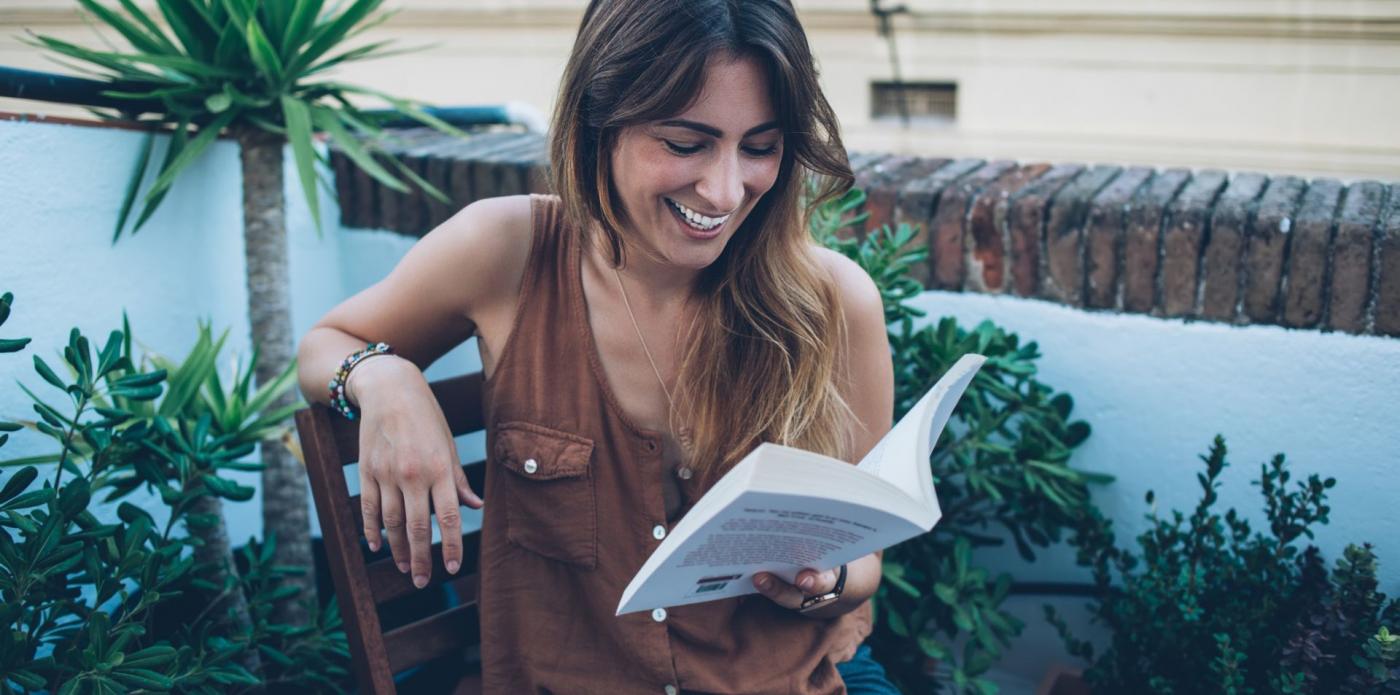 Una giovane donna seduta su una terrazza ride leggendo un libro
