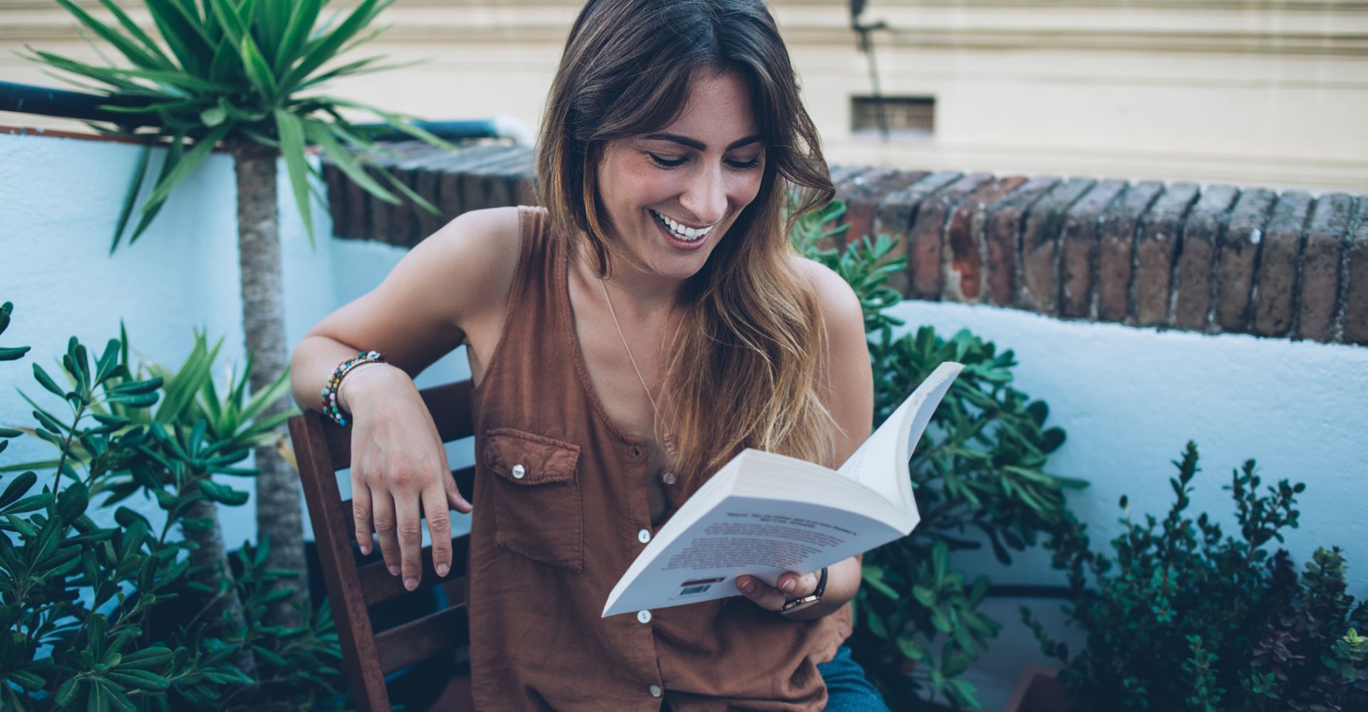 Una giovane donna seduta su una terrazza ride leggendo un libro