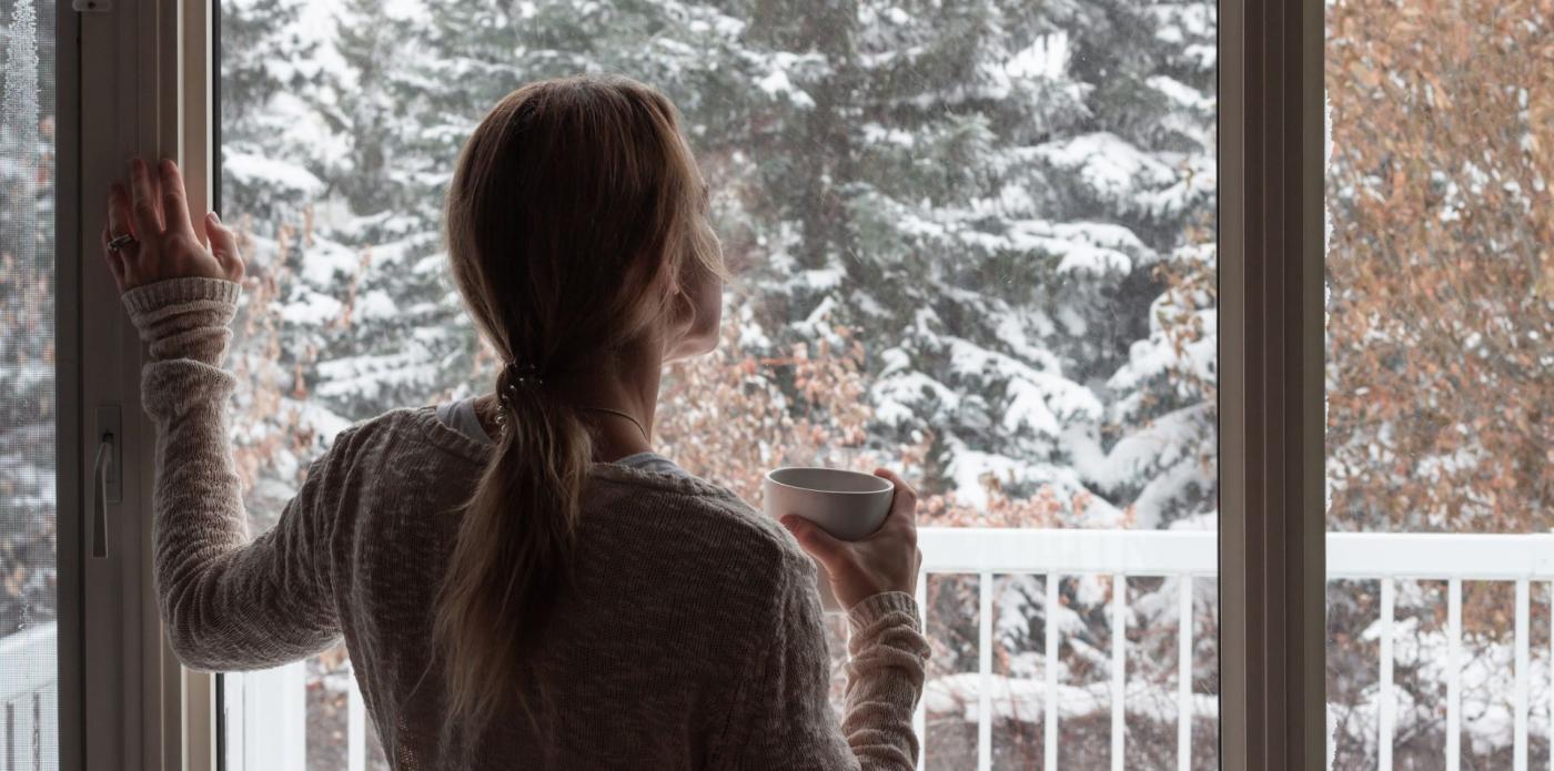 Una ragazza di spalle guarda fuori dalla finestra e sorseggia un tazza di tè durante una giornata d'inverno
