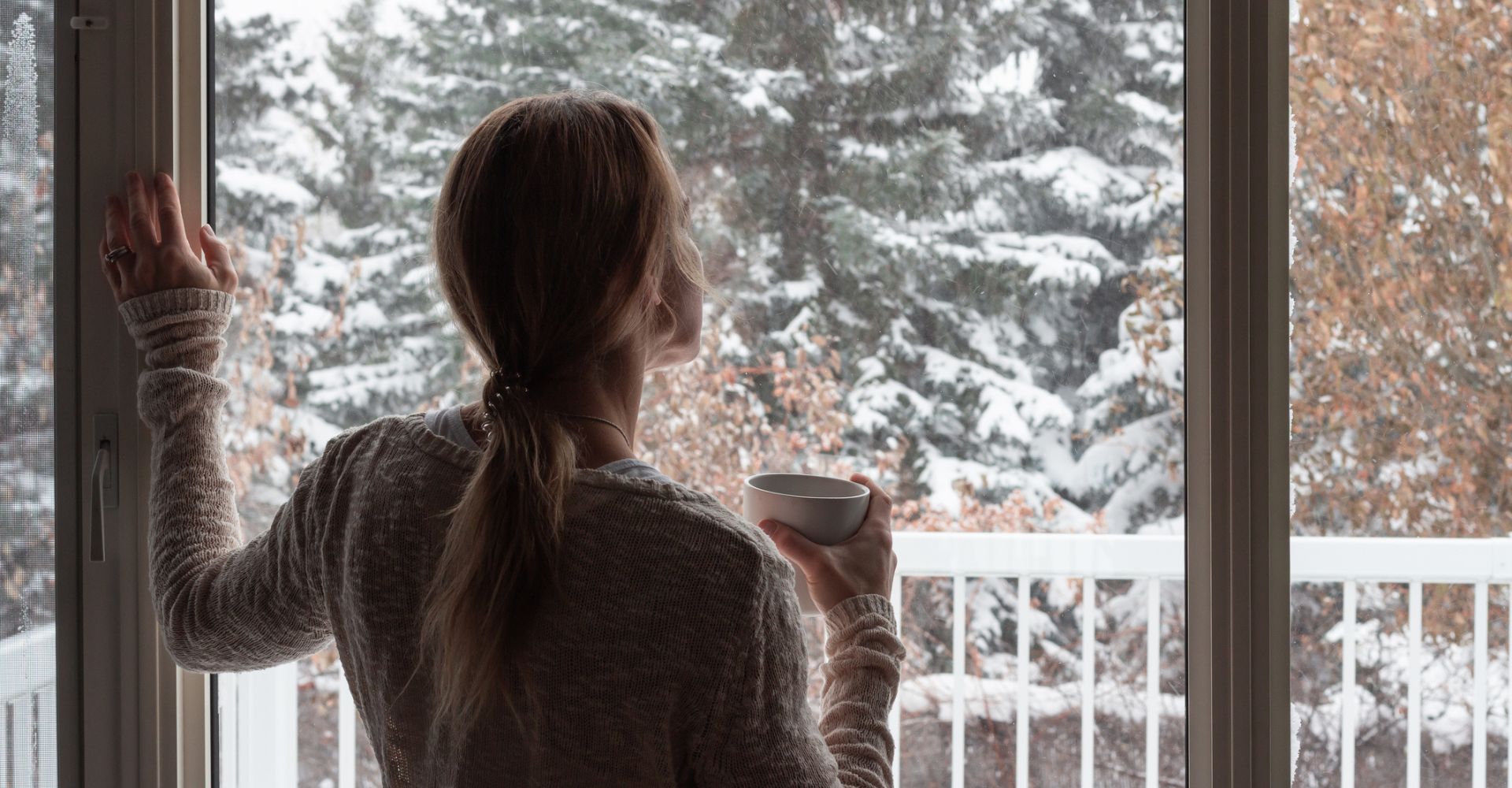 Una ragazza di spalle guarda fuori dalla finestra e sorseggia un tazza di tè durante una giornata d'inverno