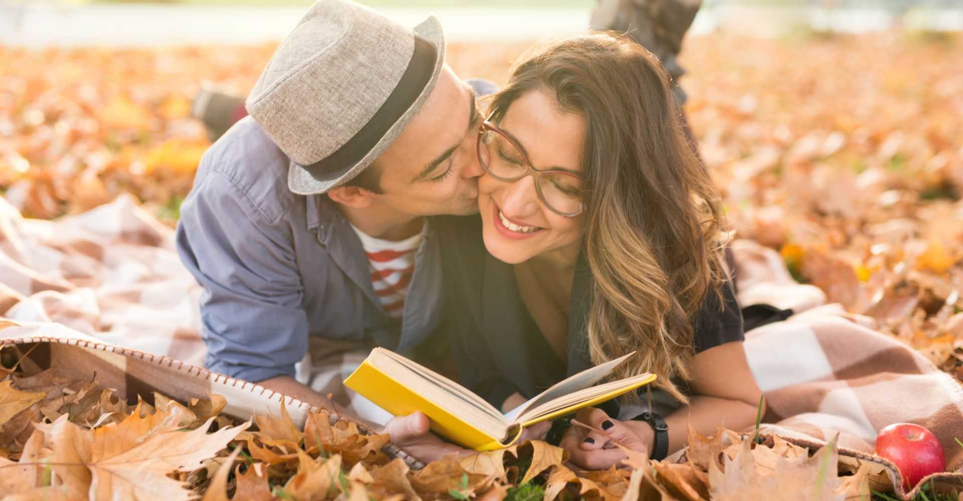 Un ragazzo sdraiato su un tappeto di foglie bacia la ragazza accanto a sé mentre entrambi tengono in mano un libro