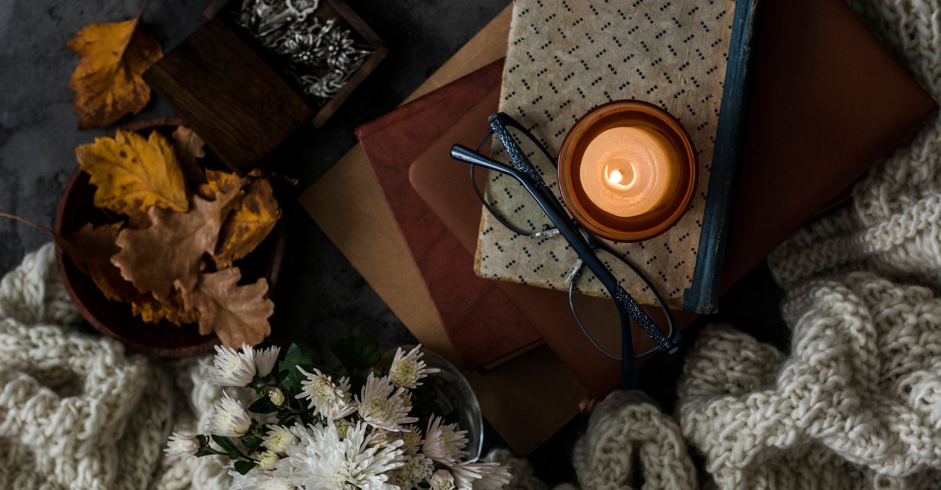 Una composizione vista dall'alto dalle atmosfere Dark Academia, con una candela accesa e un paio di occhiali poggiati sopra una pila di libri, che sono circondati da foglie autunnali e sciarpe di lana