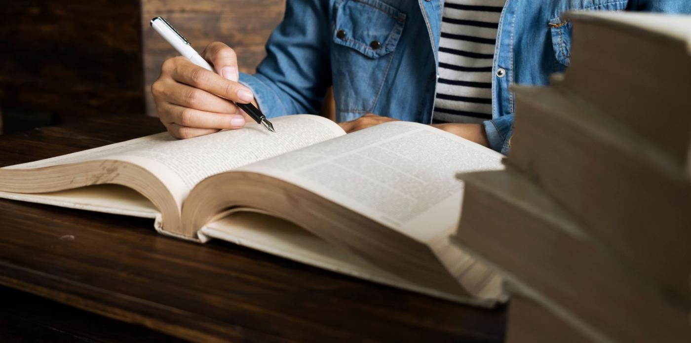 Una ragazza seduta a una scrivania consulta un dizionario mentre accanto a lei c'è una pila di altri vocabolari l'uno sull'altro