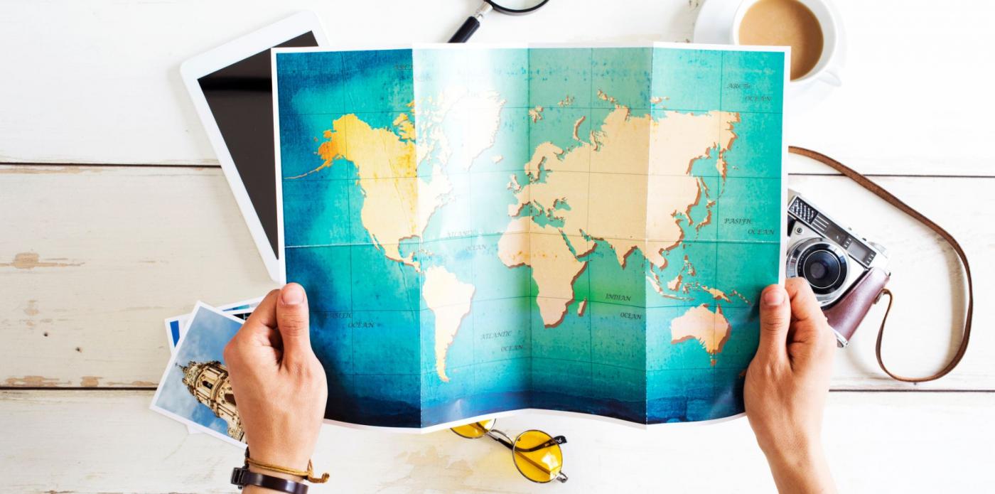 Una ragazza tiene tra le mani una mappa del mondo mentre sul tavolo davanti a lei sono poggiate una macchina fotografica, una tazza di caffè, una lente di ingrandimento e delle fotografie