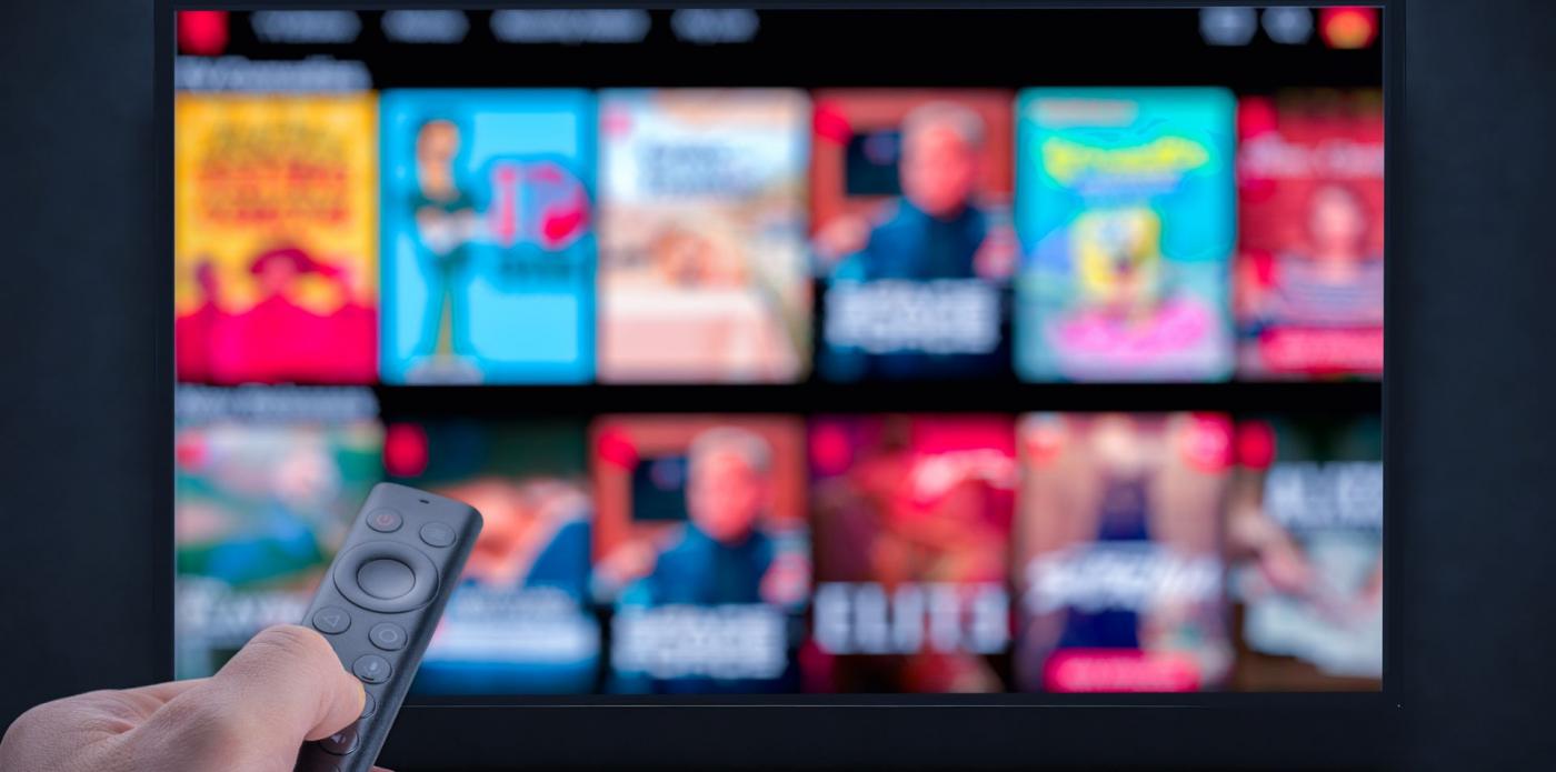 Una mano punta il telecomando verso lo schermo di una tv in cui appare l'anteprima di diversi serie tv, in correlazione alle serie tv tratte dai libri che usciranno nel 2025