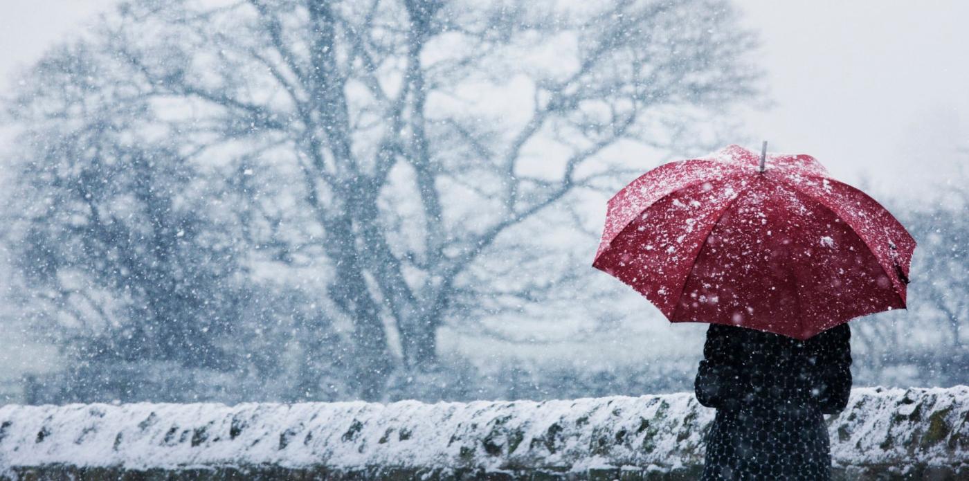 Una ragazza di spalle tiene in mano un ombrello rosso, riparandosi dalla neve che cade intorno a lei e agli alberi che la circondano