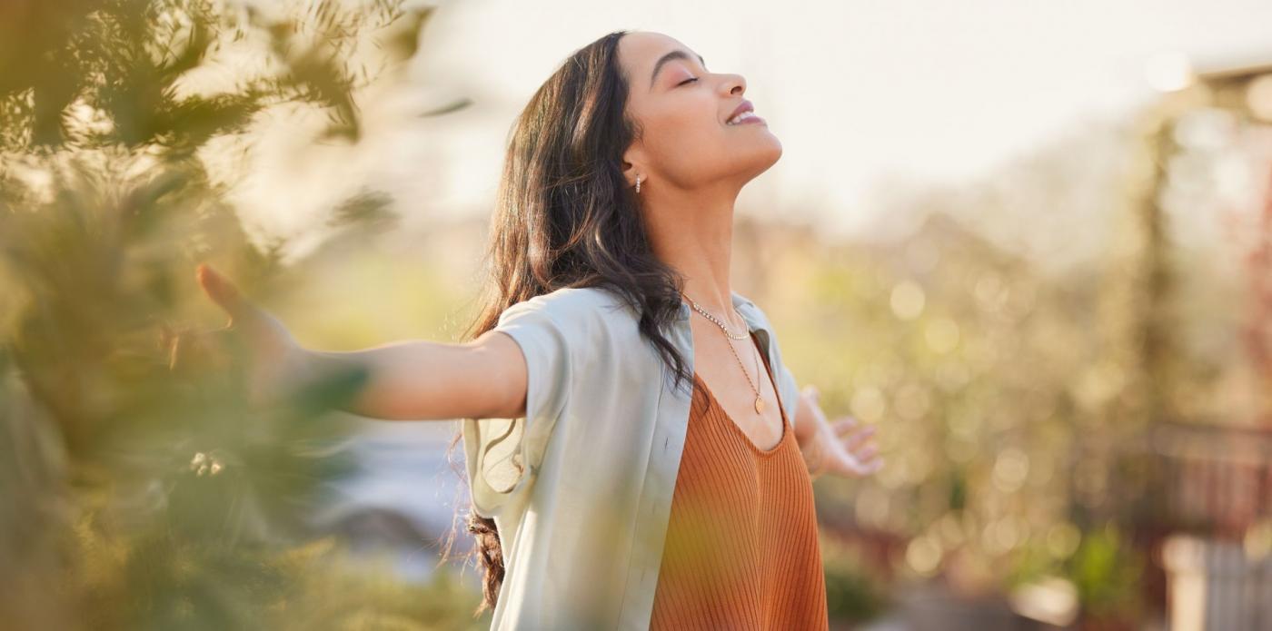 Una ragazza immersa nella natura sorride a occhi chiusi alla luce del sole, aprendo le braccia con leggerezza e serenità