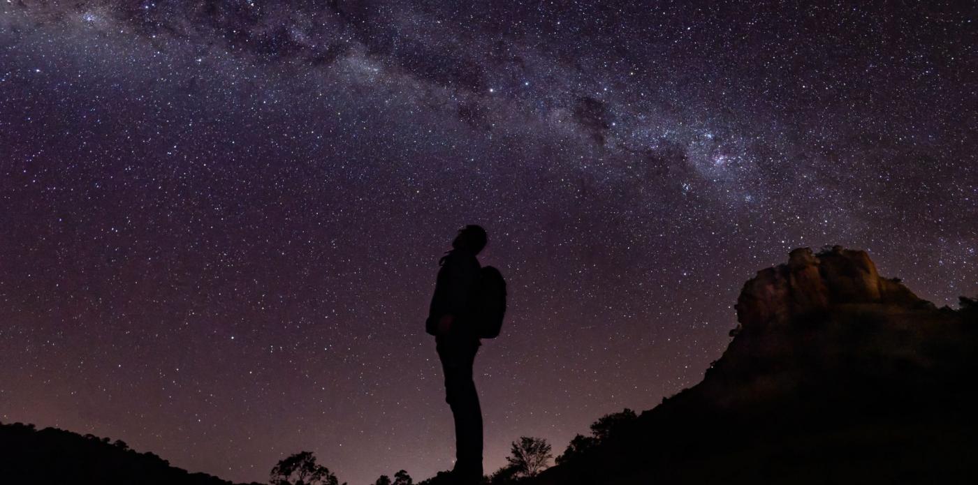 Silhouette di un ragazzo di profilo che osserva il cielo stellato da un'altura con uno zaino in spalla