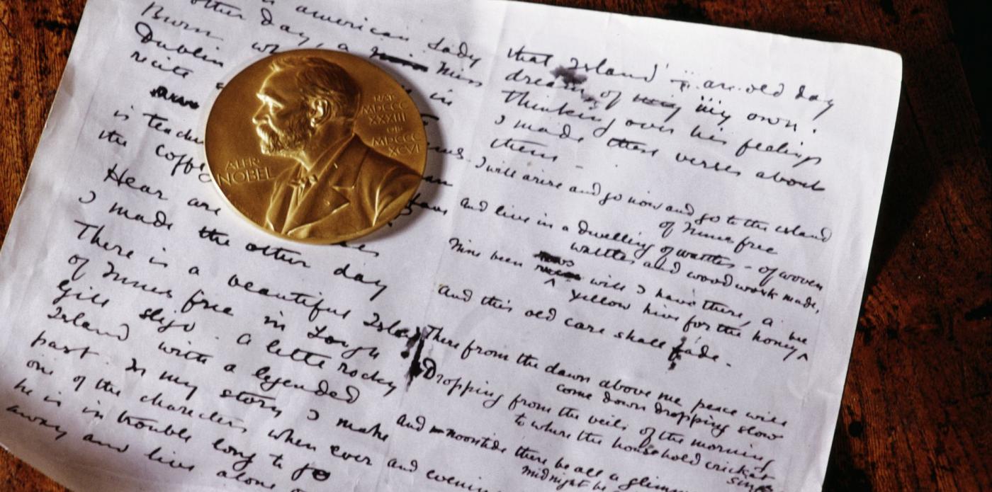Un foglio scritto a mano poggiato su un tavolo di legno, sopra il quale si trova una moneta dedicata al Premio Nobel per la Letteratura