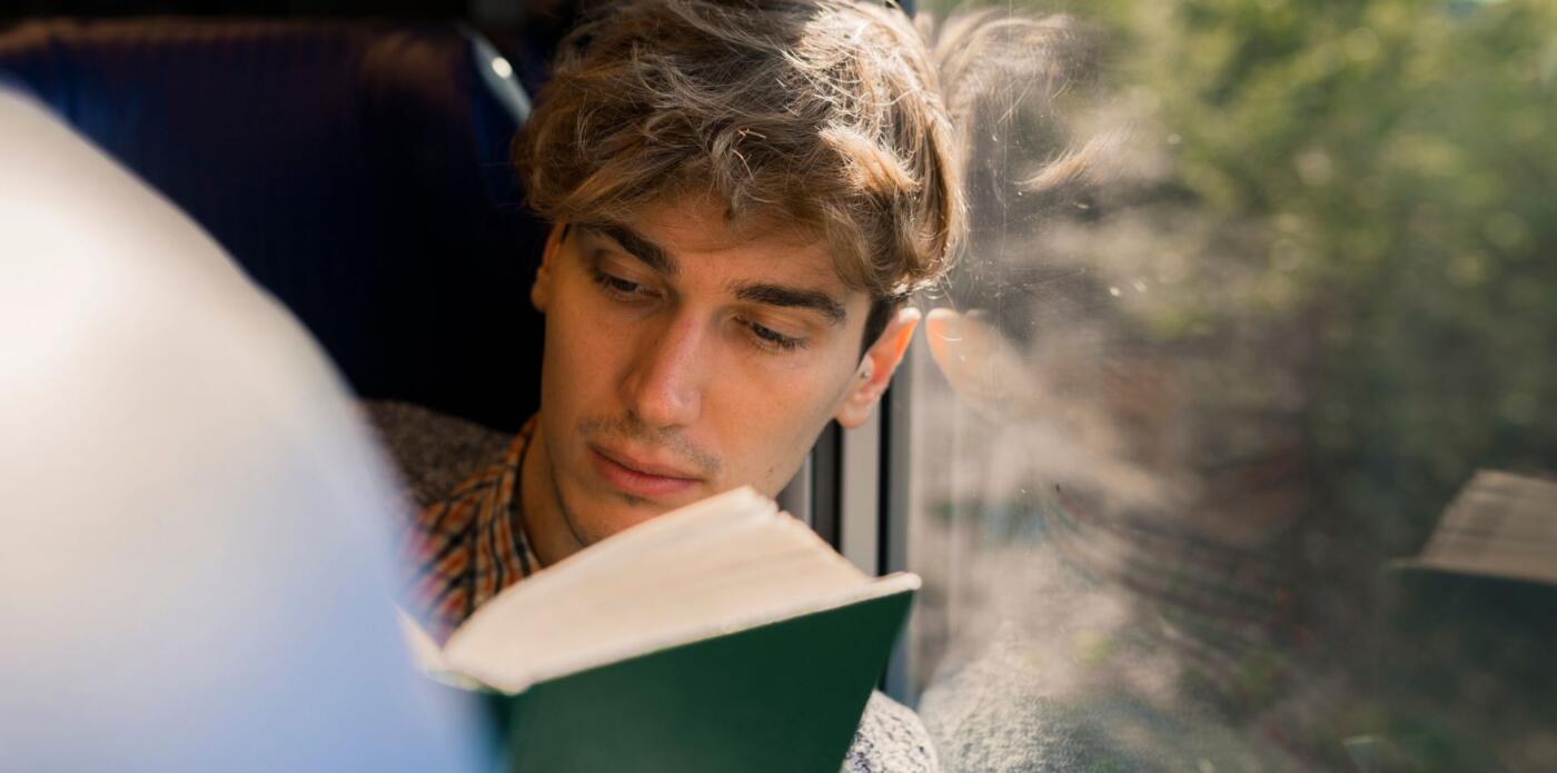 Primo piano di un ragazzo che legge un libro mentre è seduto e si appoggia al finestrino di un treno in corsa